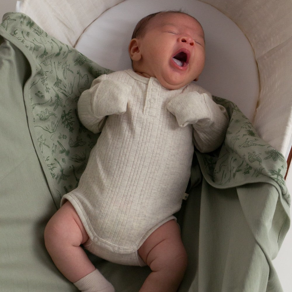 Newborn baby in a white onesie yawning in a crib with green bedding.