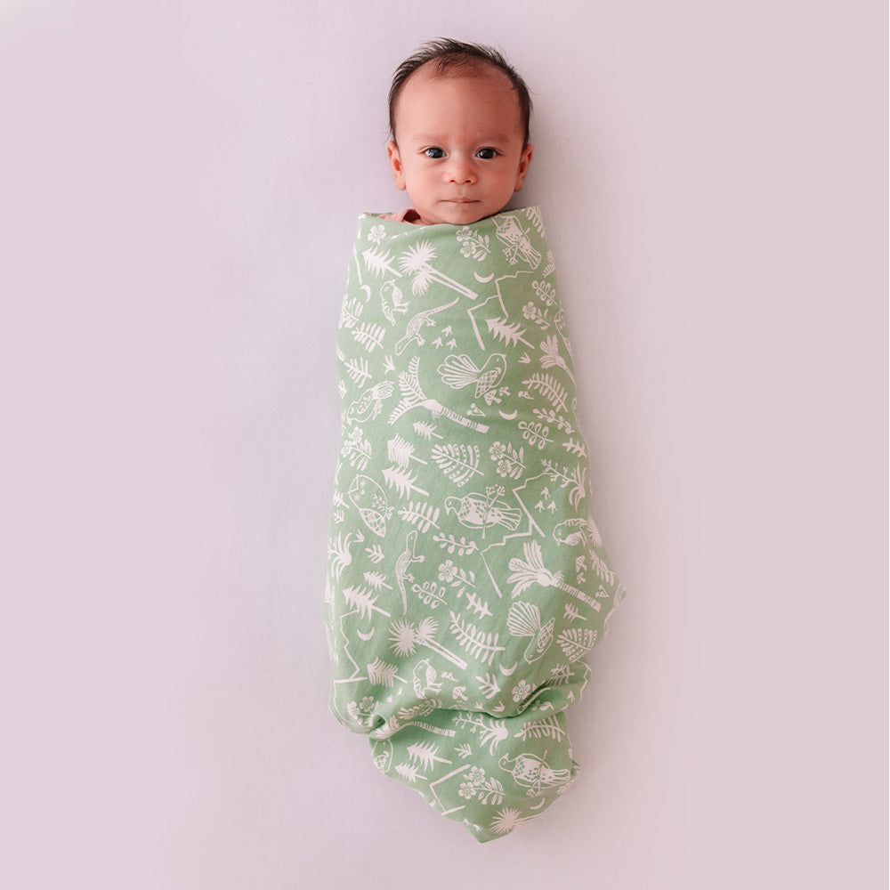 Baby swaddled in a green blanket with white patterns on a light purple background