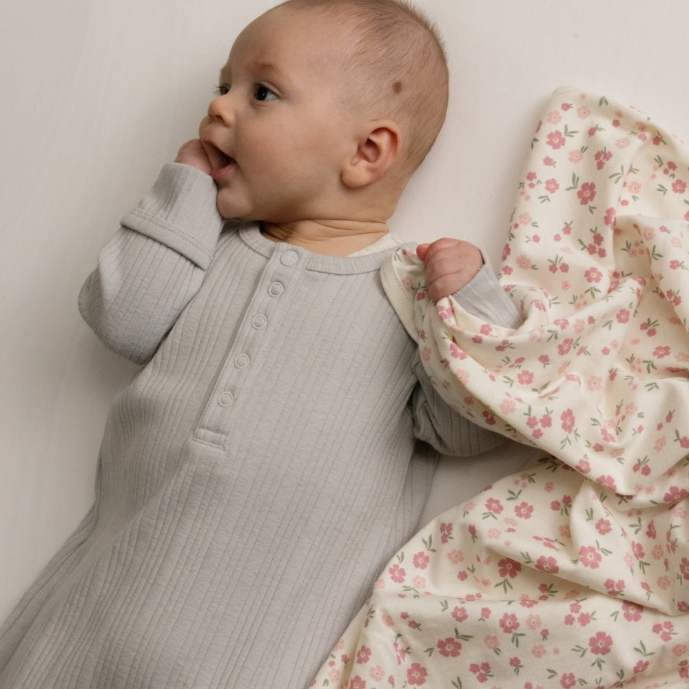 A baby wearing a light gray ribbed long-sleeve henley gown, lying on a white surface next to a cream-colored floral blanket with pink flowers.