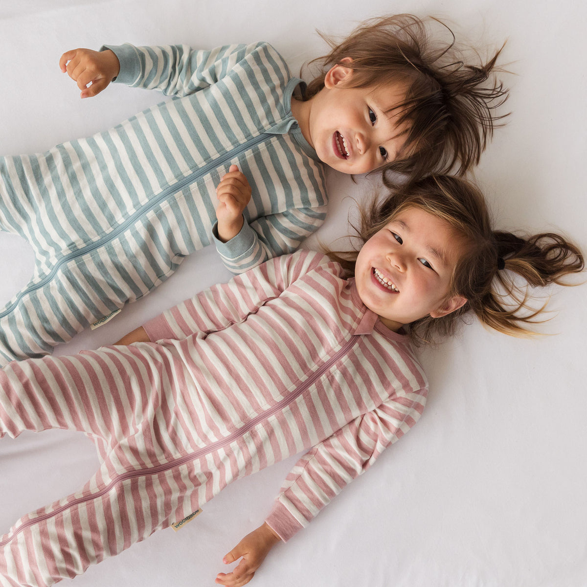 Two children lying on a white sheet wearing striped woolbabe PJ Suits