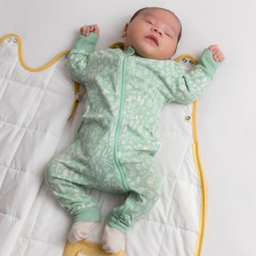 Baby in a green patterned onesie lying on a white blanket with yellow straps.