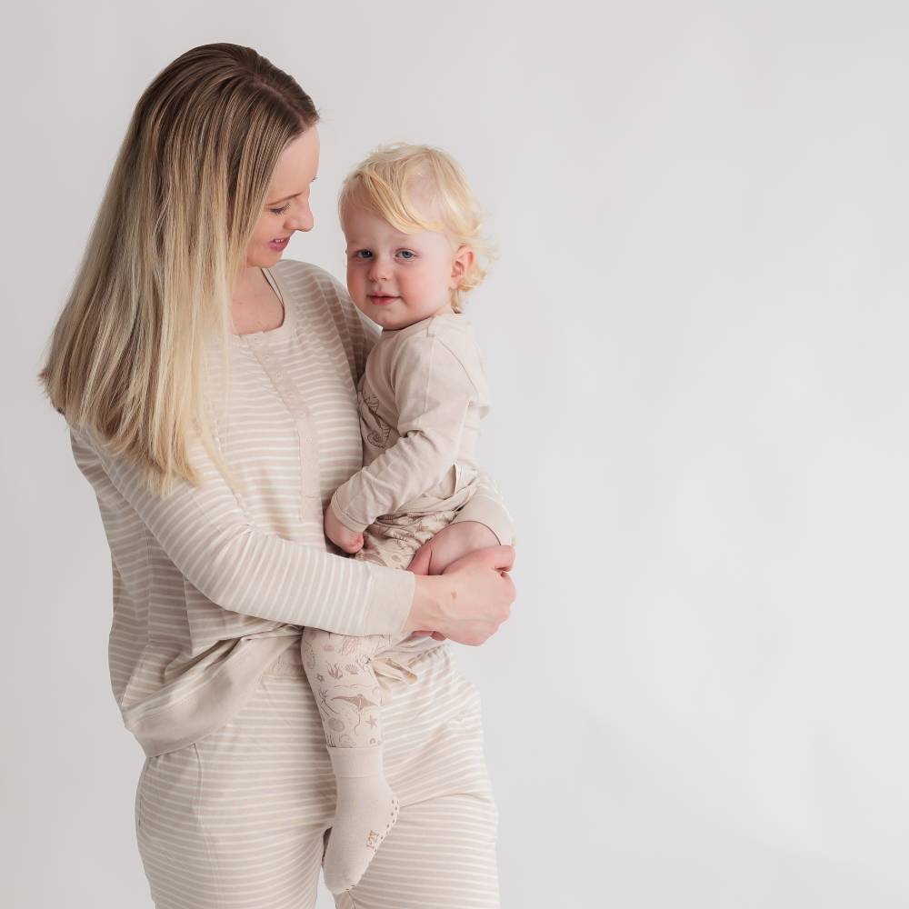 Woman holding a baby in matching beige outfits against a plain background