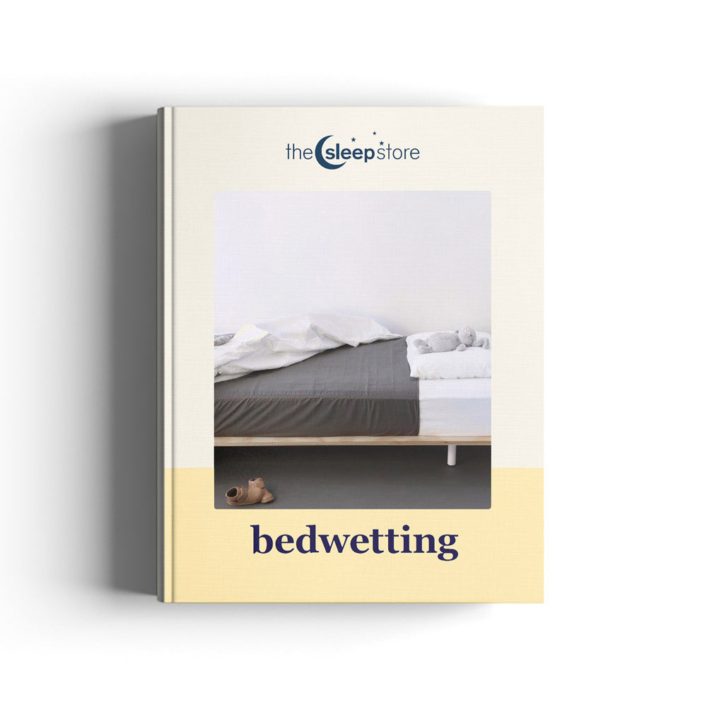 An ebook titled 'bedwetting' by 'the sleep store' is shown standing upright against a white background. The cover features a photograph of a bed with gray bedding and a small teddy bear on the floor, with the title 'bedwetting' printed in dark blue text on a pale yellow band at the bottom.