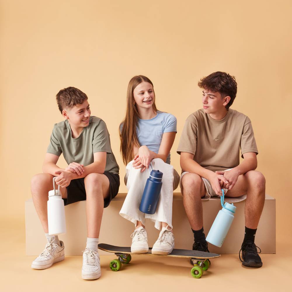 Three young people sitting on a beige block with skateboards and water bottles, against a beige background