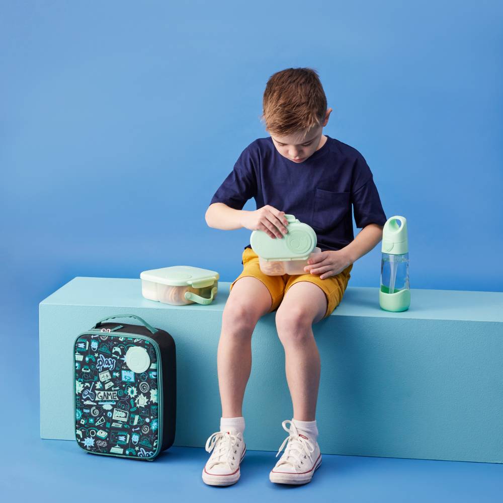 Child sitting on a blue bench with b.box lunch supplies including an insulated lunch box and water bottle against a blue background.