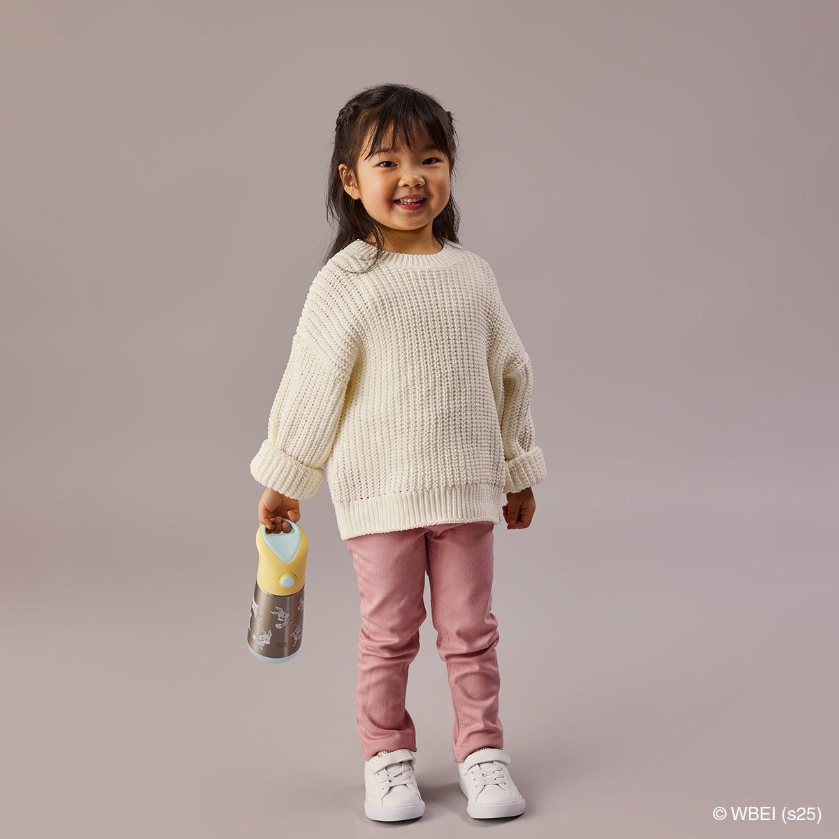 Child wearing a cream sweater and pink pants holding a Looney Toons bottle against a plain background