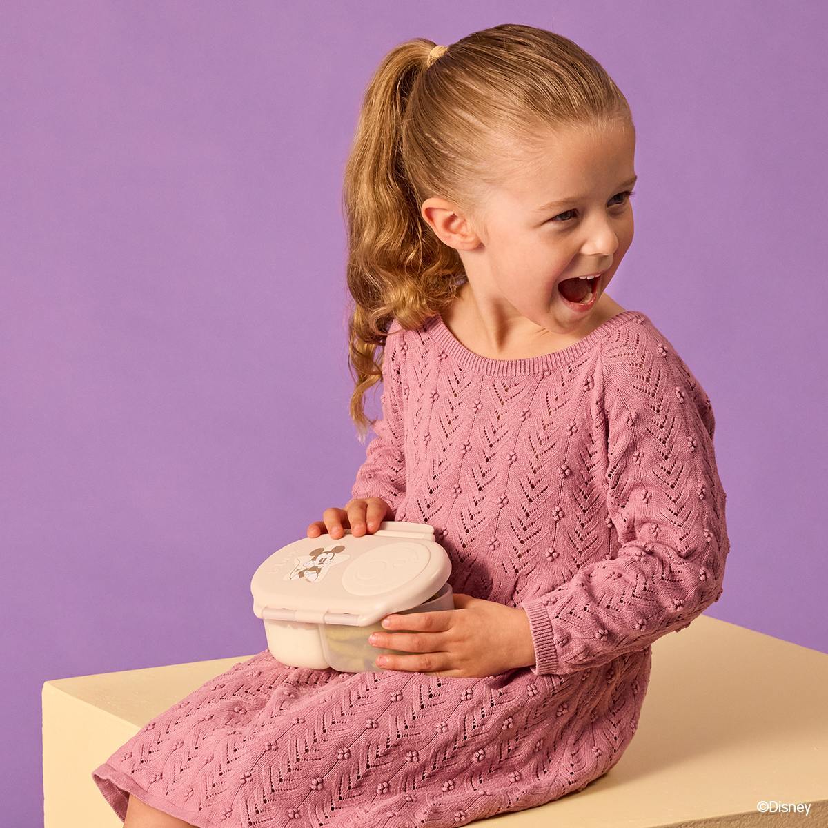 A young child sitting down smiling, about to open a beige snackbox. 