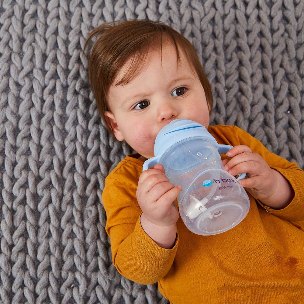 Baby in a mustard yellow outfit drinking from a blue sippy cup on a textured gray blanket.