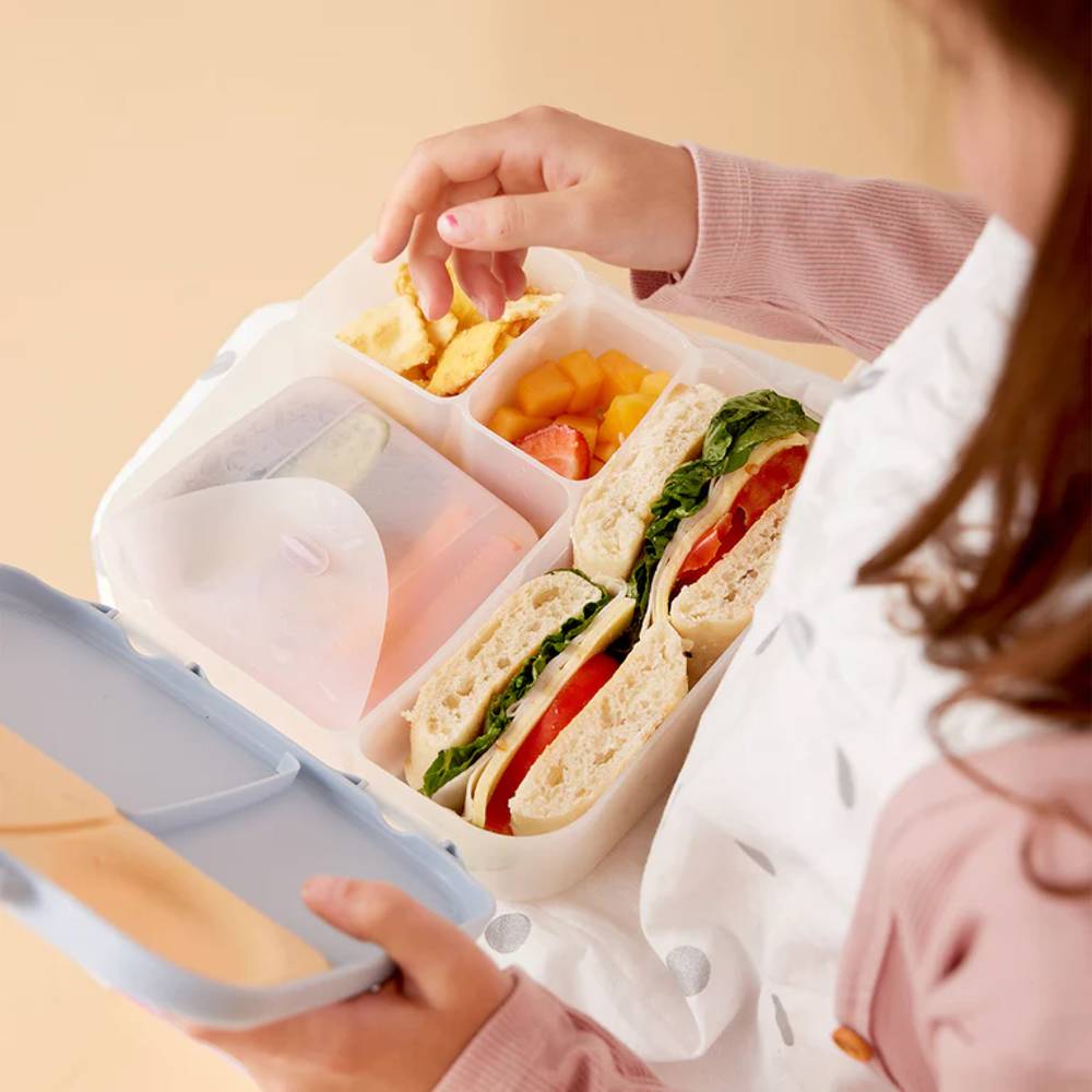 Person opening a bento box with compartments containing a sandwich, fruit, and other items.