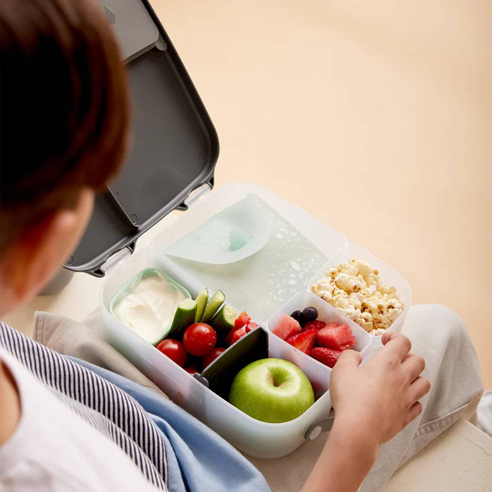 Person opening a bento box with compartments containing fruits, vegetables, and popcorn.