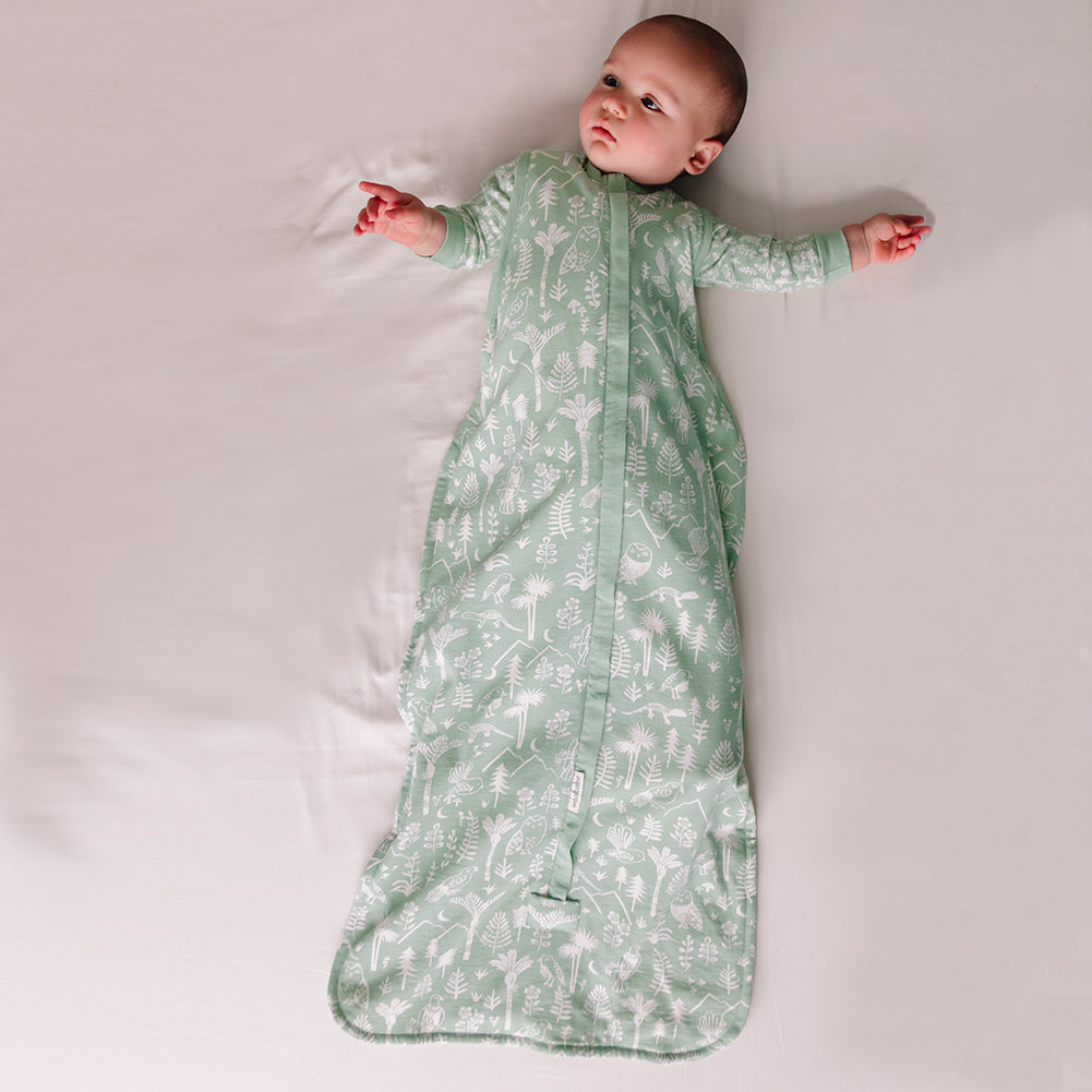 Baby lying on a white surface wearing a green patterned wilderness print sleeping bag sack