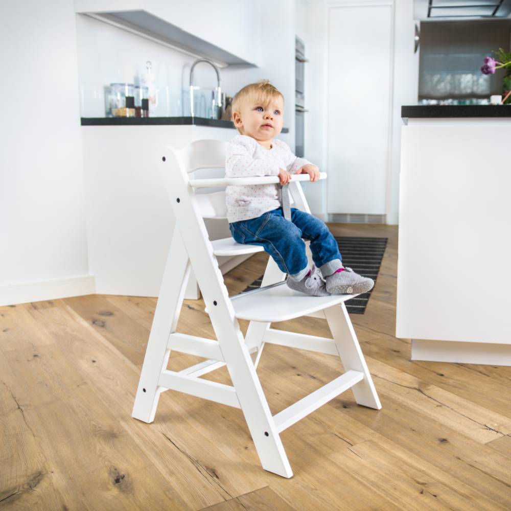 Child sitting on a white wooden adjustable chair in a modern kitchen.