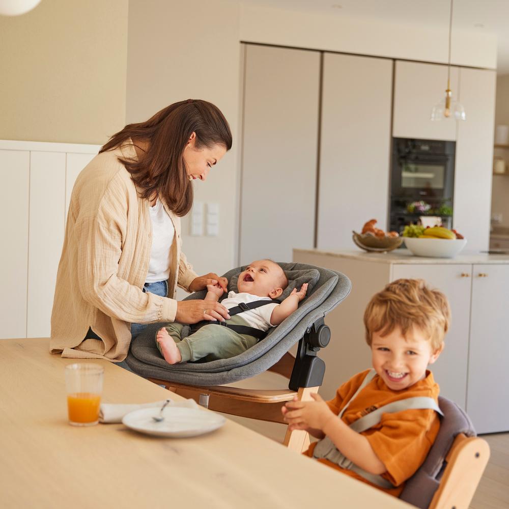 Woman with a baby in a high chair and a child sitting next to her in a different high chair in a kitchen.
