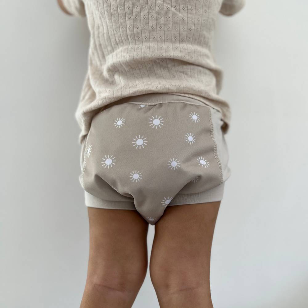 Child wearing beige training pants with sun pattern and beige top on a plain background