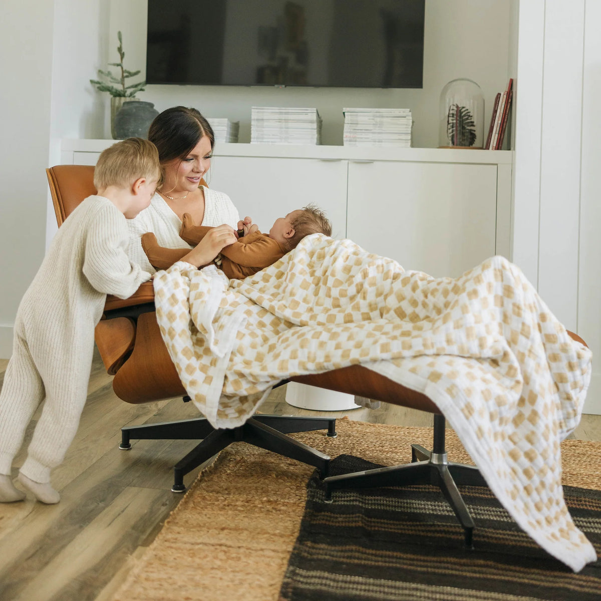 Woman holding a baby wrapped in a beige and white check patterned little unicorn throw blanket in a living room setting and toddler beside her.