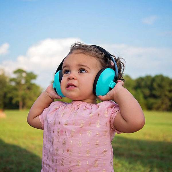 A toddler girl wearing bright turquoise over-ear headphones and a pink patterned dress stands outdoors in a grassy field under a blue sky.
