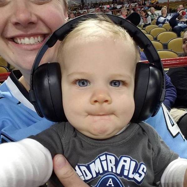 Baby wearing large black earmuffs with a person holding them, in a stadium setting.