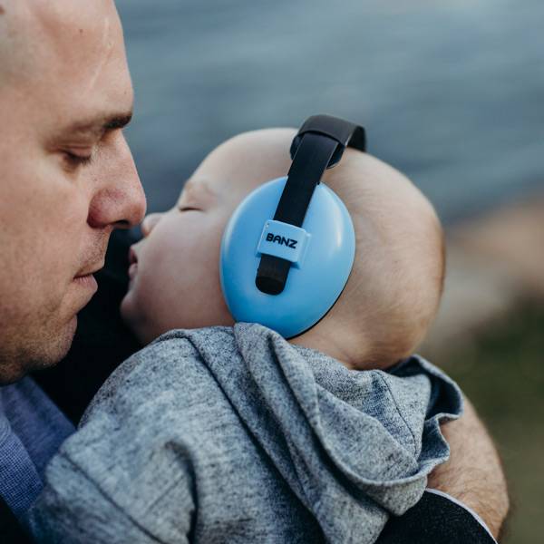 Man holding a baby wearing blue ear muffs with 'BANZ' branding, outdoors.