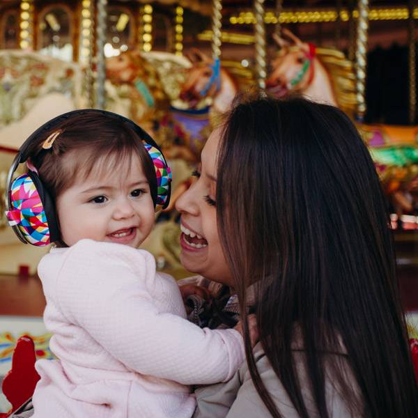 Woman holding a child with colorful earmuffs in front of a carousel.