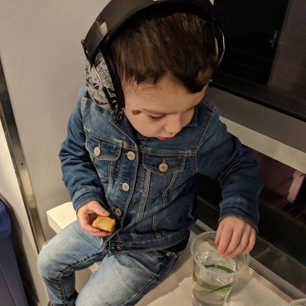 A young child wearing a blue denim jacket and jeans, sitting down on a train and wearing a black and grey over-ear earmuffs. The child is holding a piece of food and reaching towards a clear plastic cup on the table.