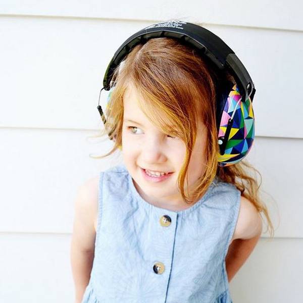 A young girl with light brown hair smiling while wearing large over-ear earmuffs with a colorful geometric pattern on the earmuff, set against a white paneled wall.
