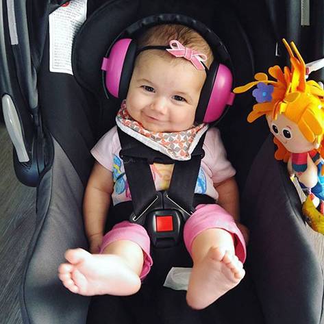 A baby in a capsule car seat with pink earmuffs and a plush toy to the right 