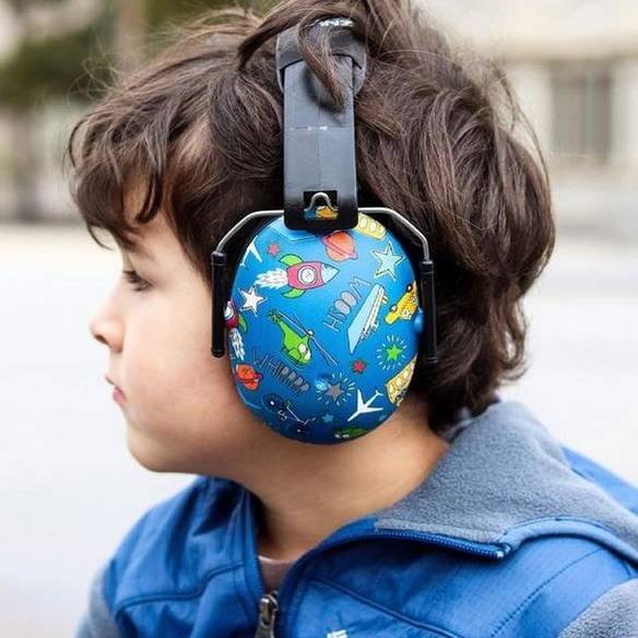 A child in profile wearing blue over-ear headphones with a colorful space-themed print of rockets, planets, and stars, set against a blurred outdoor background.