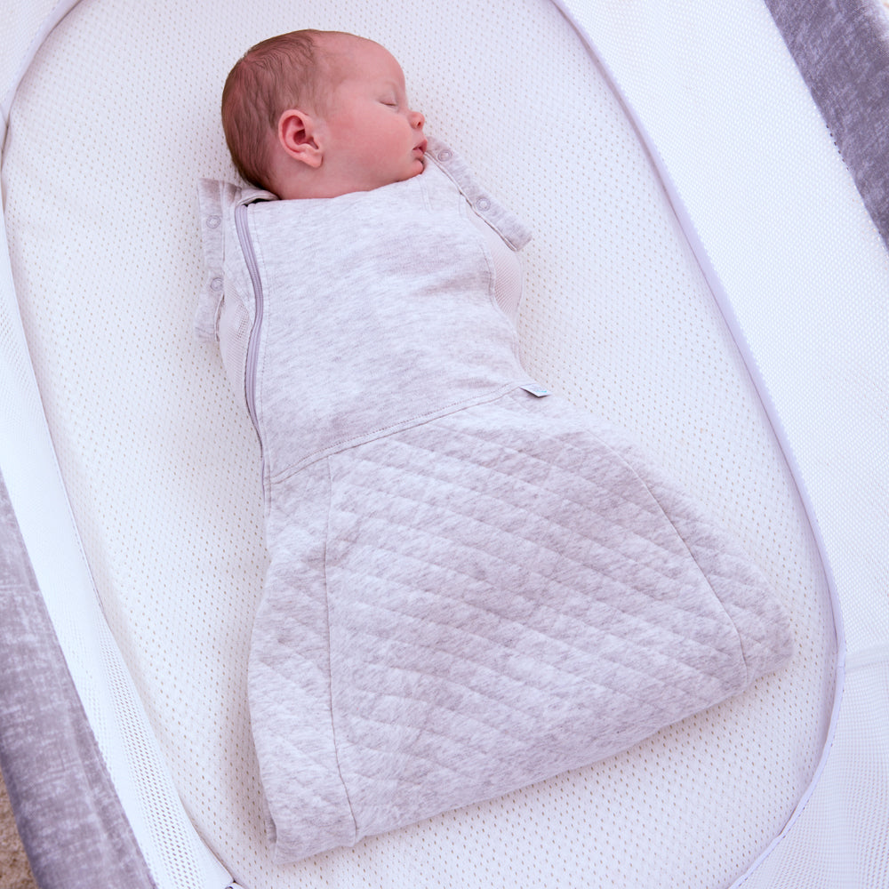 Newborn baby swaddled in a light gray quilted swaddle sleeping bag lying on a white crib.