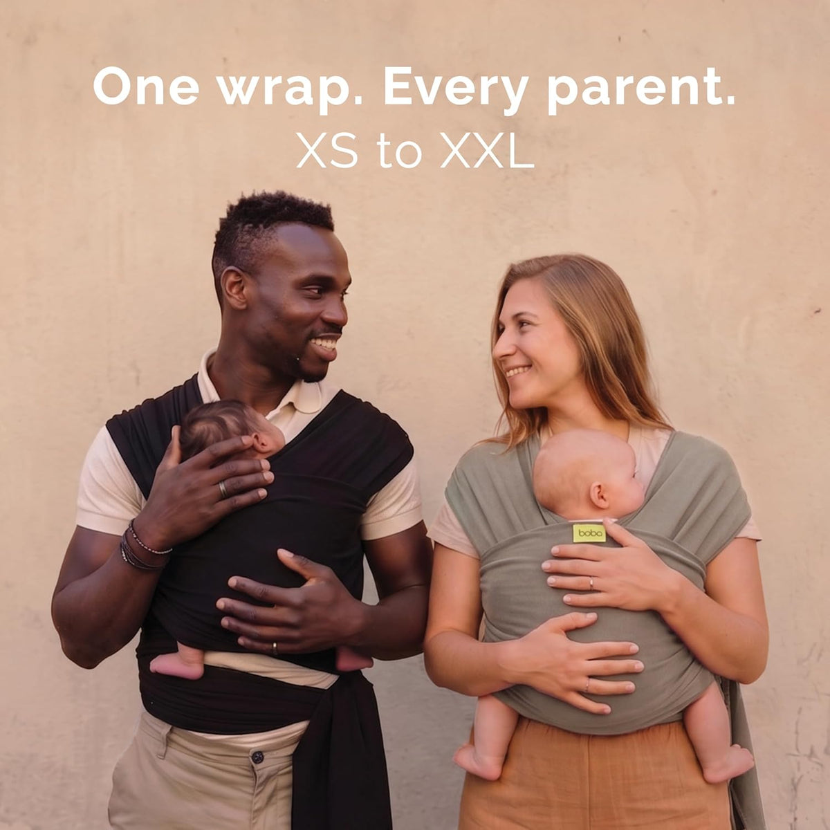 A man and a woman wearing baby carrier wraps, each holding a baby. The man's wrap is black and the woman's wrap is a muted sage green.