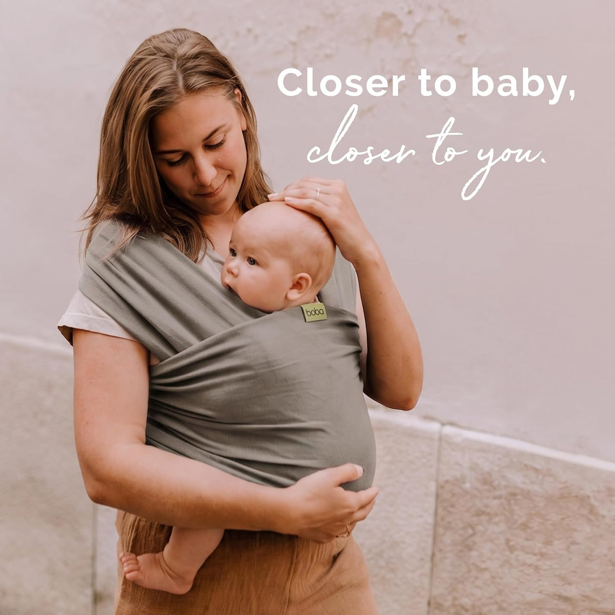 A woman wearing a gray baby wrap carrier holds a baby against her chest. The wrap has a small green OHO logo patch.
