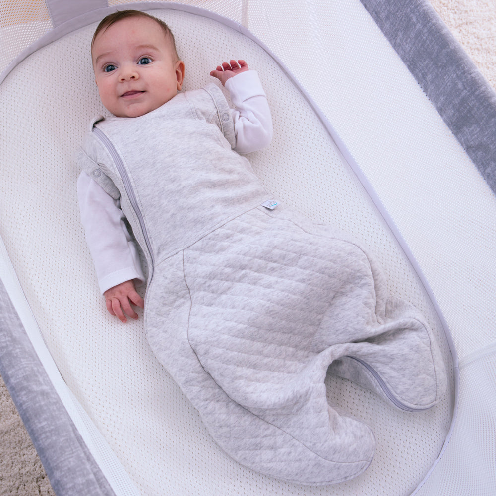 Newborn baby swaddled in a light gray quilted swaddle sleeping bag with arms out lying on a white crib.