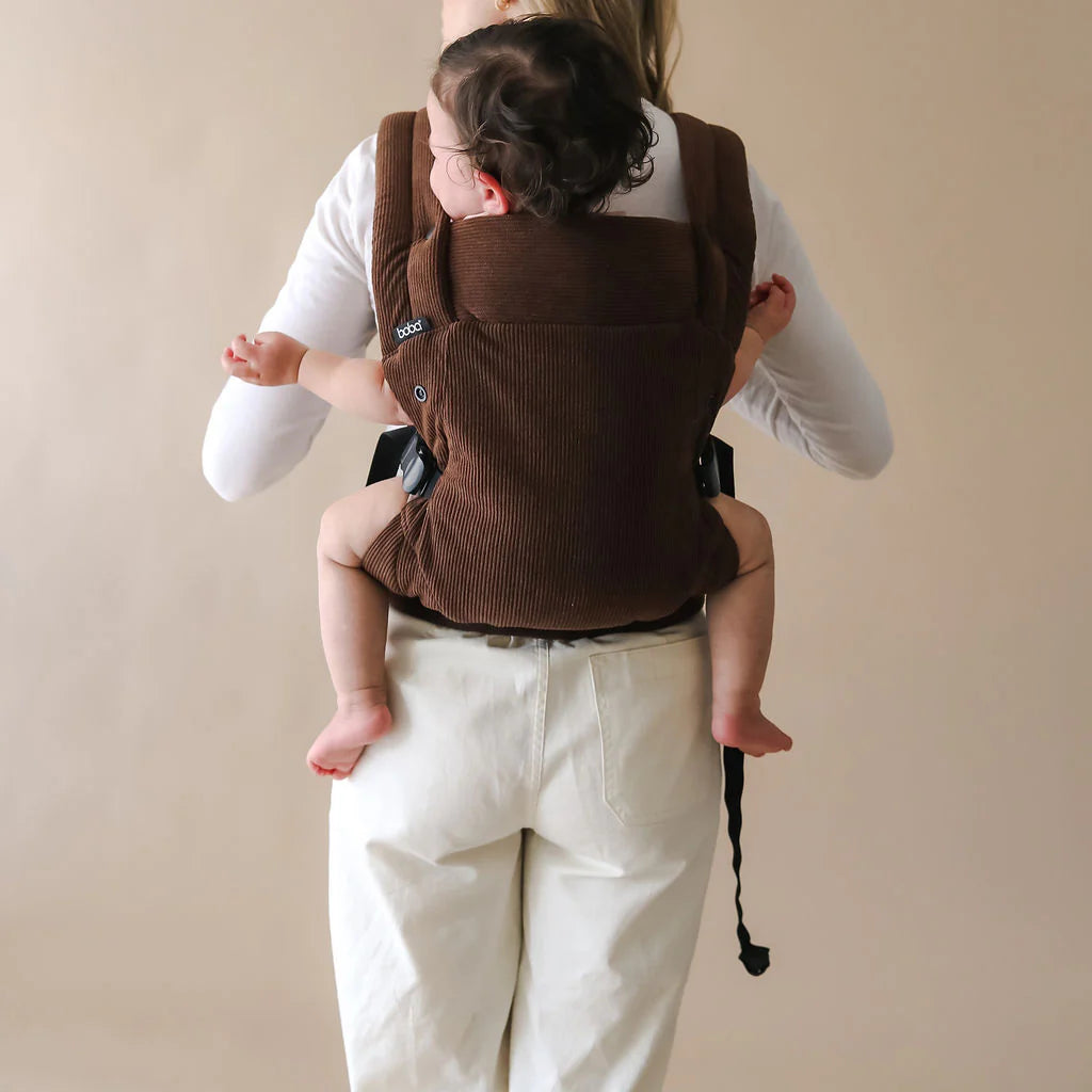 Person wearing a baby in a brown carrier in the back carrying position against a beige background