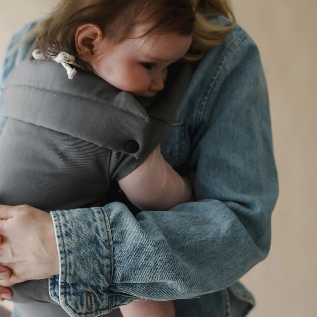 A baby carrier worn con an adult, shown in a close-up lifestyle shot. The carrier is made of a soft, solid gray fabric and features a black snap button closure.