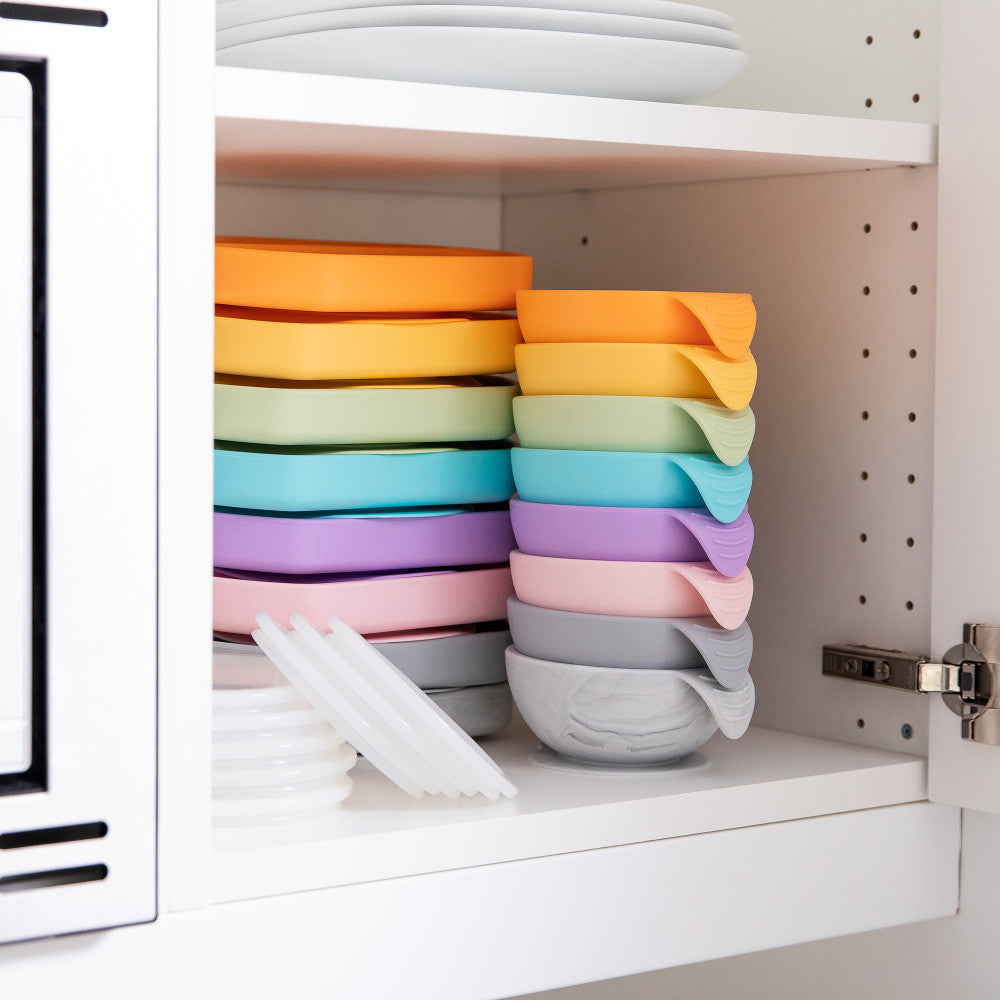 A stack of different coloured Bumkins silicone bowls and plates with the clear lids sitting next to them inside a white cupboard 
