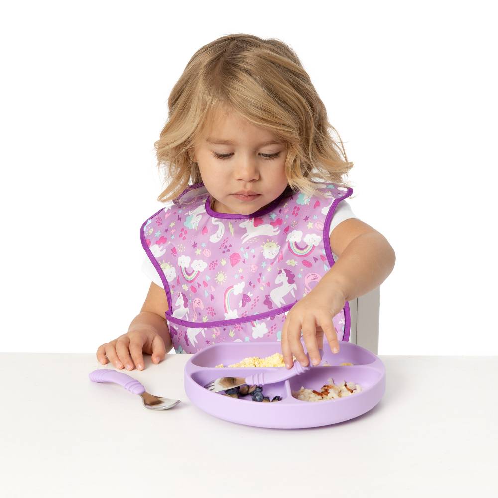 A blonde child wearing a purple unicorn bib using bumkins lavender cutlery to eat from a lilac purple grip dish with food inside 