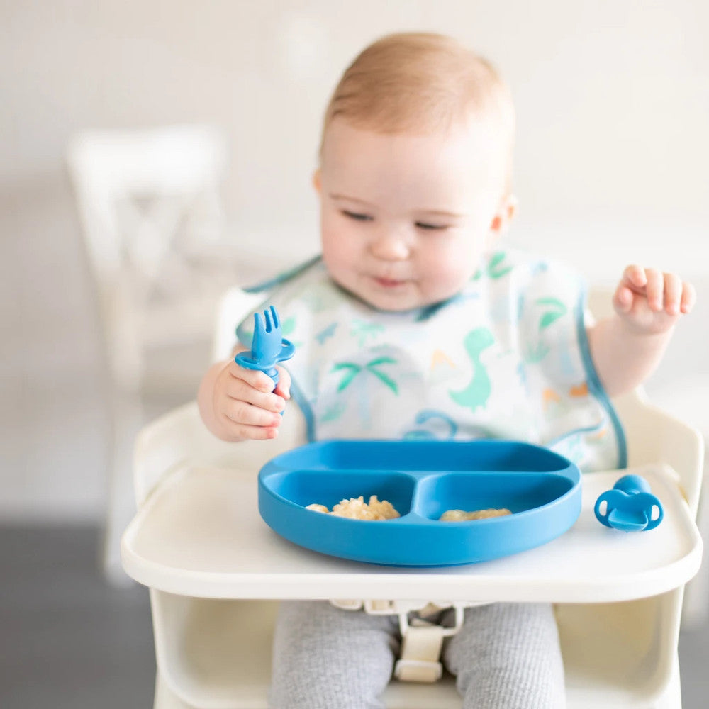 A light haired child in a blue dino bib and grey pants sits in a white highchair holding a chewtensil cutlery fork. A dark blue bumkins grip dish is attached to the tray with food inside.