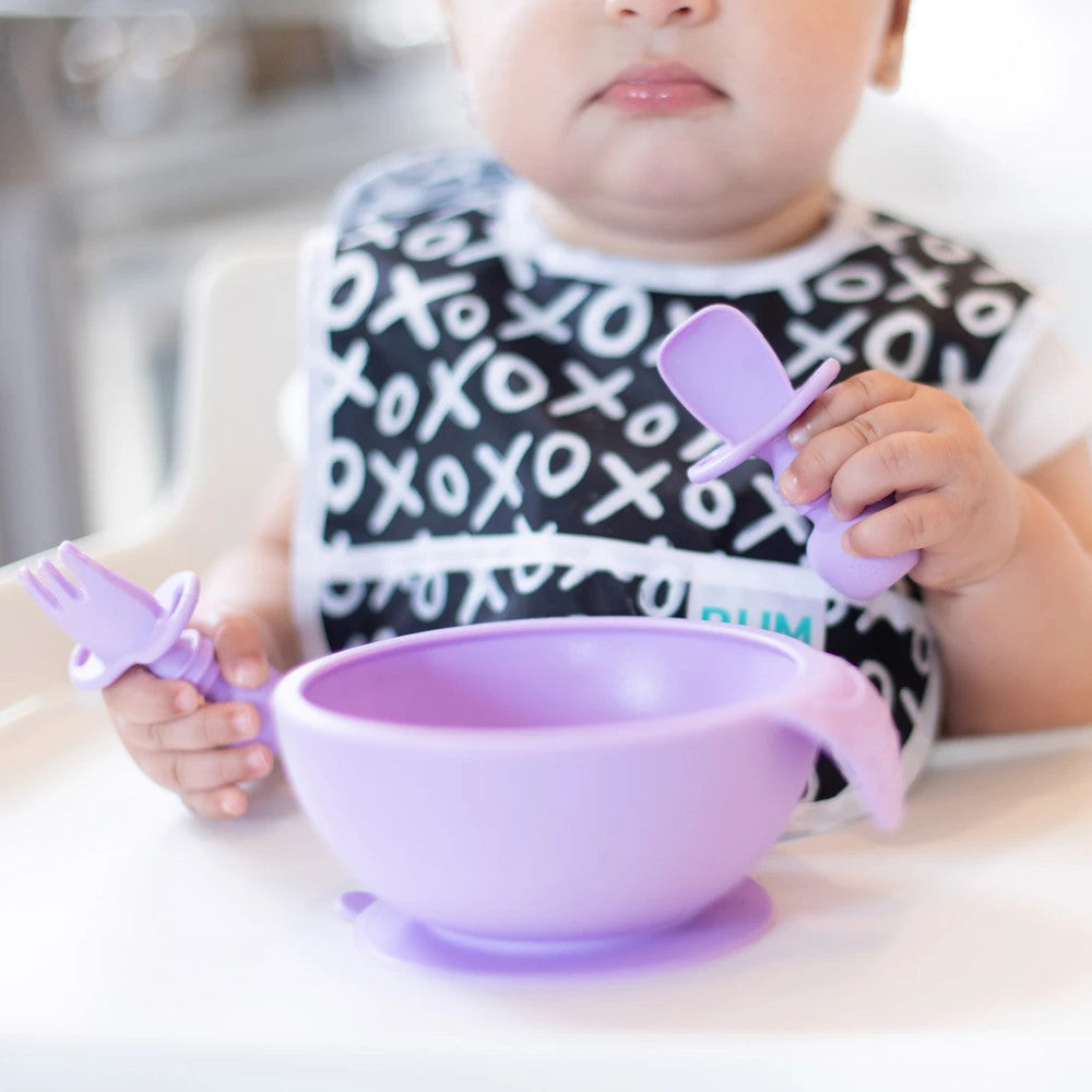 A close up of a baby sitting ina white highchair wearing a black and white bib holding a lavender set of chewstensils and a purple silicone bumkins feeding bowl stuck to the tray