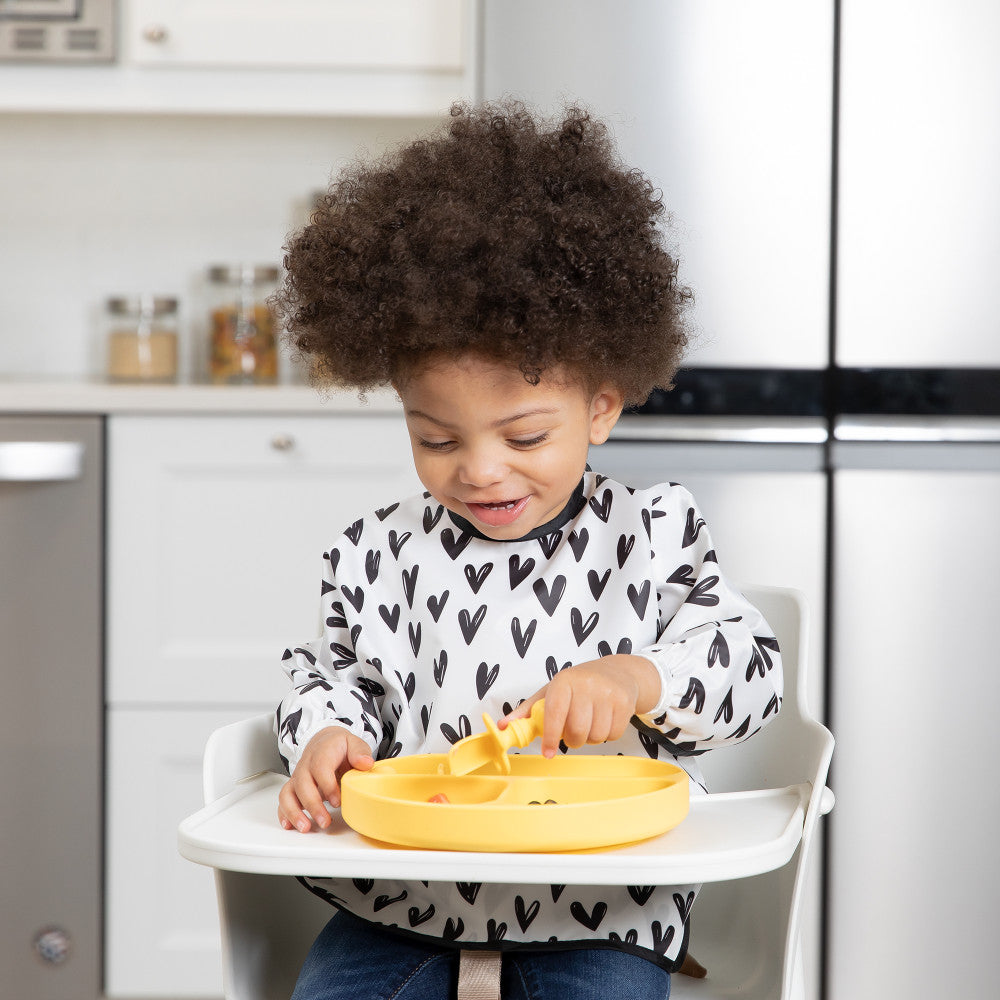 A child with dark curly hair in a black and white heart sleeved bib sits in a  white highchair holding a yellow chewtensil cutlery spoon. A yellow bumkins grip dish is attached to the tray with food inside.
 