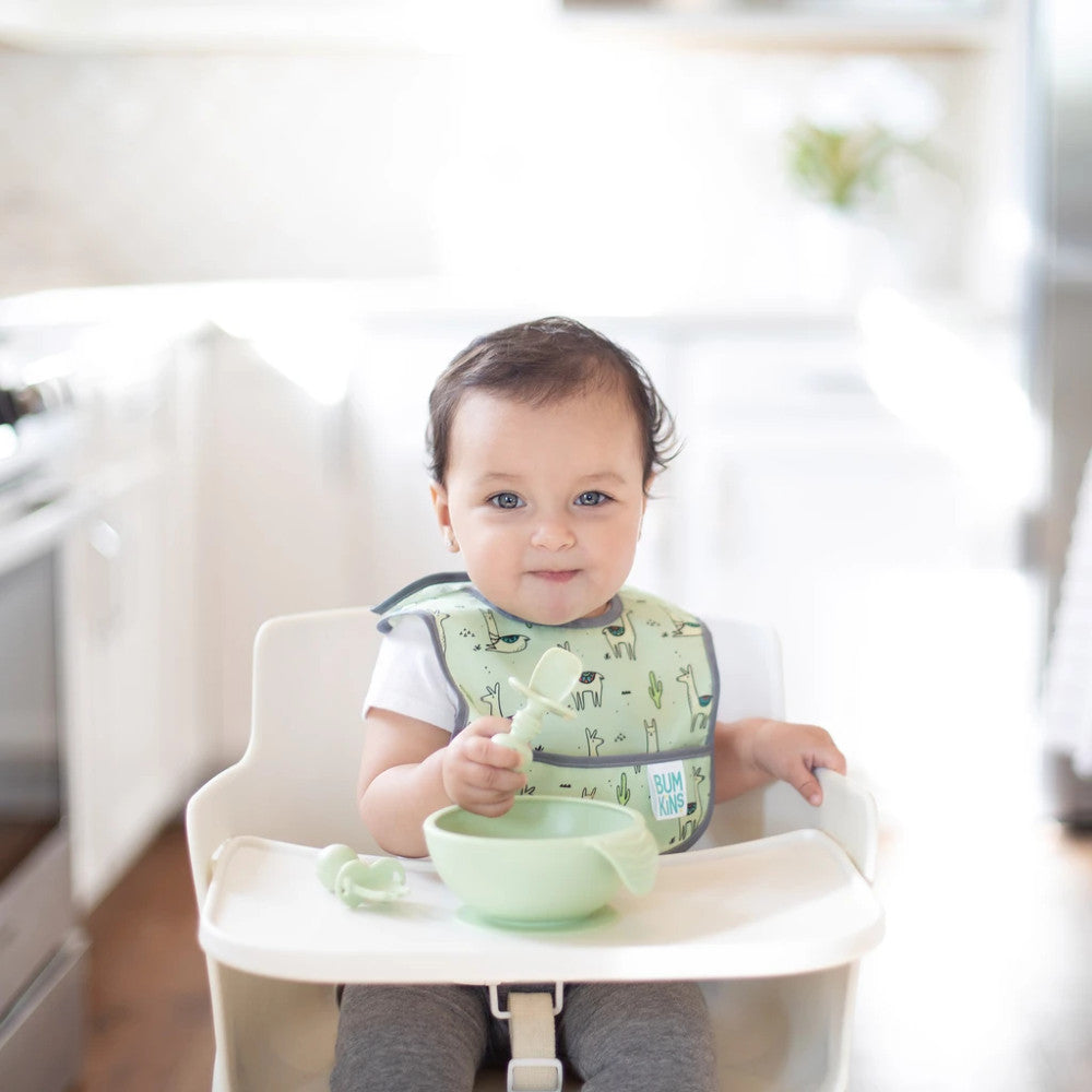 A child with dark hair in a light green bib and white shirt sits in a white highchair holding a sage chewtensil cutlery spoon. A sage bumkins feeding bowl is attached to the tray and sage silicone fork beside. 
 