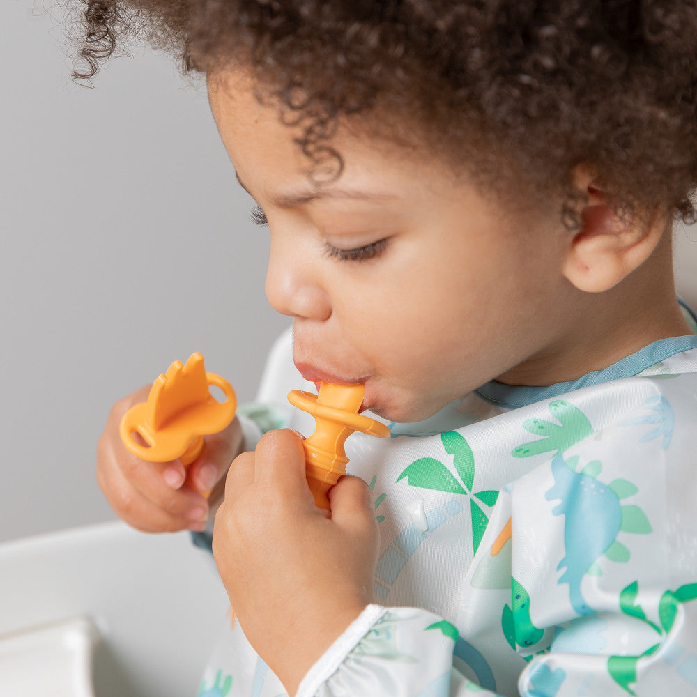 A close up of a child with dark, curly hair wearing a blue dinosaur bib using the bumkins tangerine chewtensil sppon and fork to eat their food 