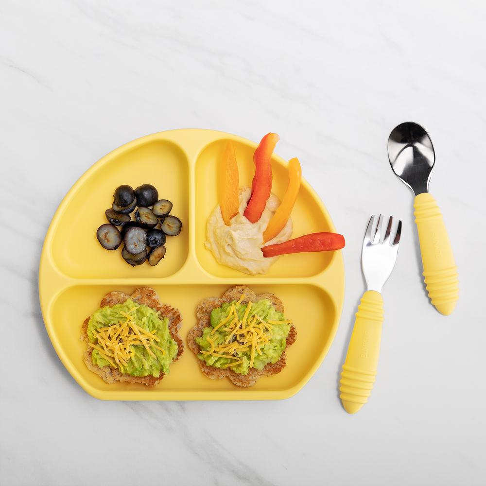 Yellow bumkins silicone grip dish with assortment of food inside next to a spoon and fork set of stainless steel cutlery with yellow silicone handles on a marble background