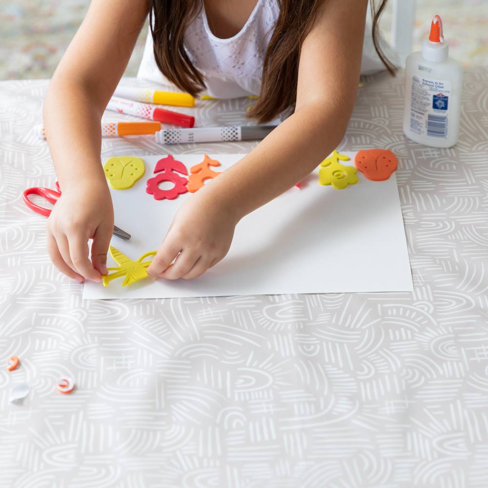 A beige bumkins splat mat with white line patterns being used for crafting by a child with dark hair. Paper, pens, scissors and glue is included 