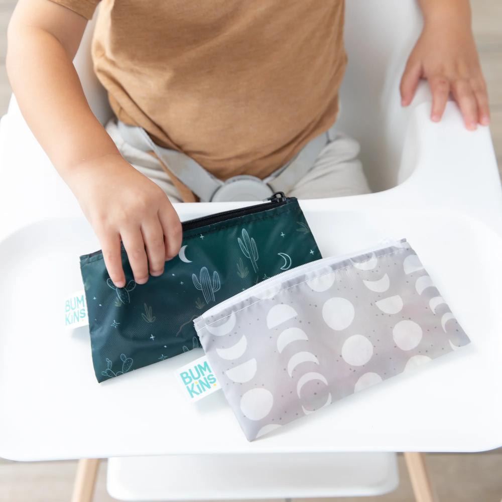 A close-up of two rectangle snack bags on a white highchair tray with a child picking up a dark green snack bag. A grey snack bag with white moon phase pattern can be found next to the green bumkins bag.