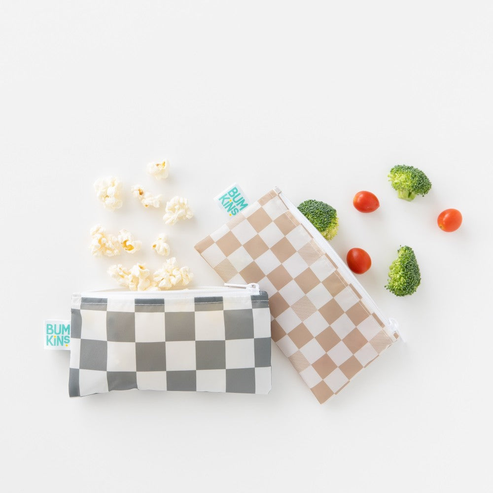 Two rectangle snack bags with white zippers. One with grey check and popcorn spilling out and one with beige and cream check with broccoli and cherry tomatoes spilling out on a white background 