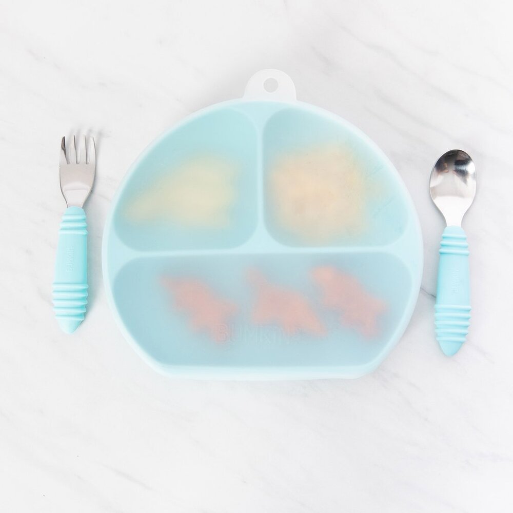 A clear stretch silicone lid shown on a blue 3-section silicone grip dish with fork and spoon located on either side on a marble background 