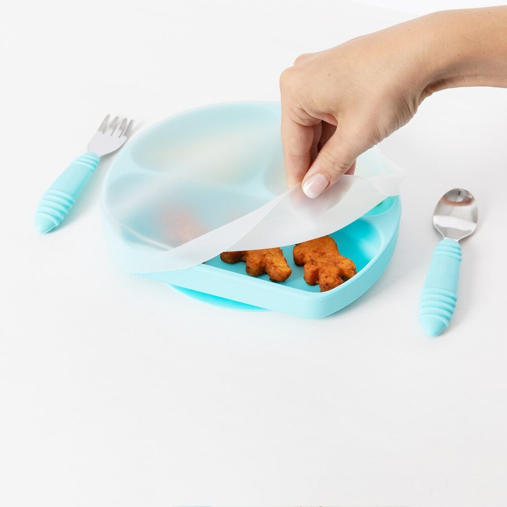 A hand lifting the corner of a clear silicone stretch lid over a blue grip dish with food inside. A blue fork and spoon a located either side of the dish on a white background 
