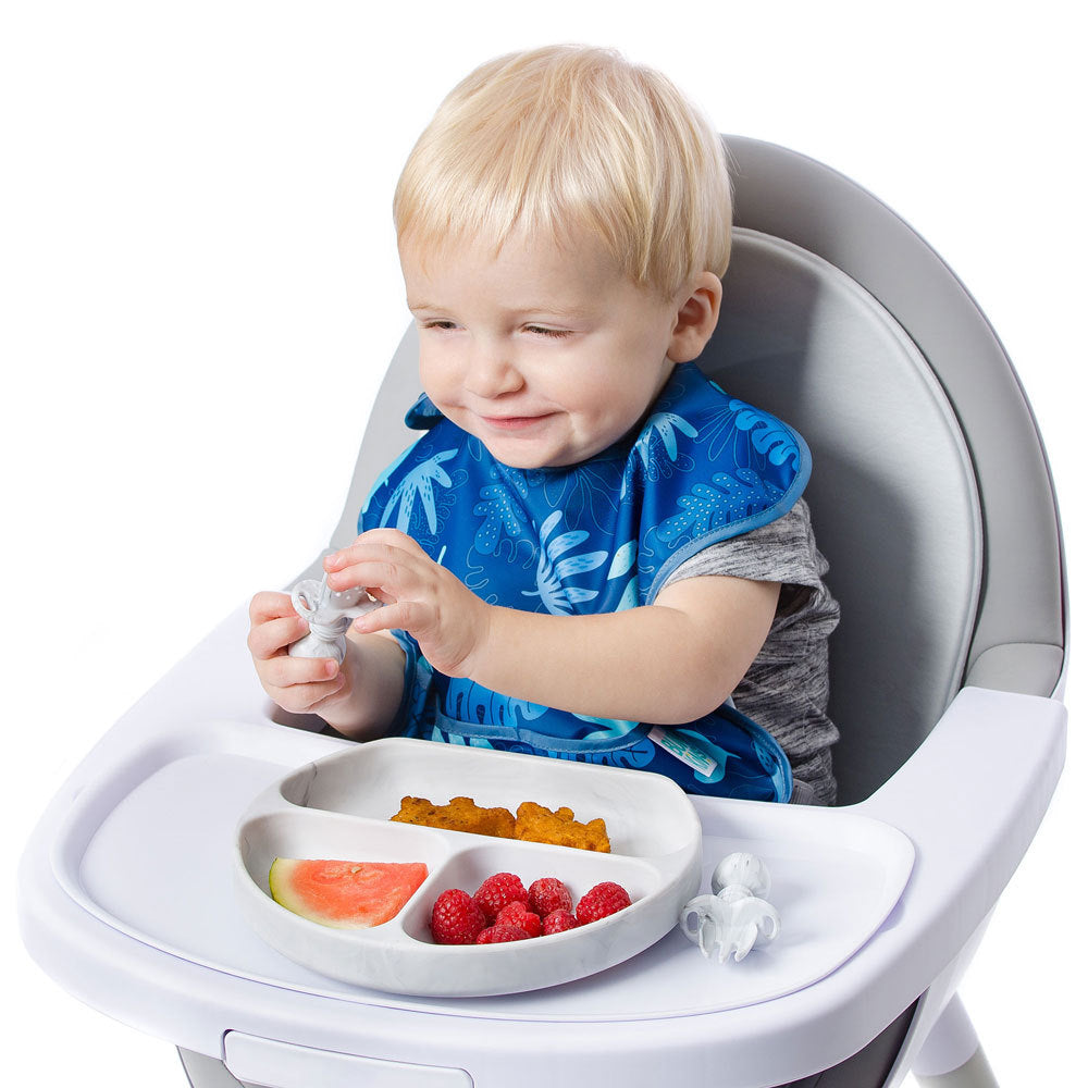 A blonde child in a blue bib and grey shirt sits in a grey and white highchair holding a chewtensil cutlery spoon. A marble bumkins grip dish is attached to the tray with food inside. 