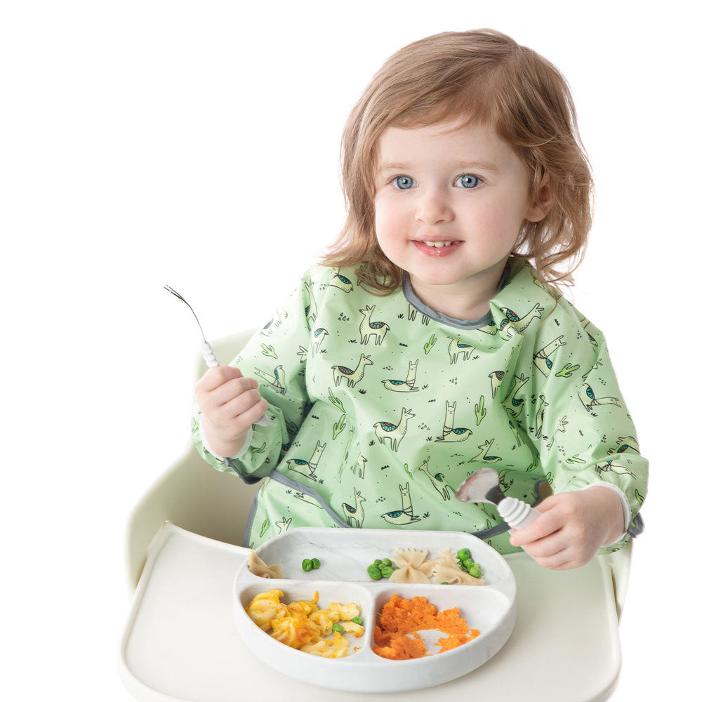 A child in a white highchair wearing a mint green sleeved bib with Llama pattern. Child is holding marble cutlery and eating from a marble grip dish with food inside 