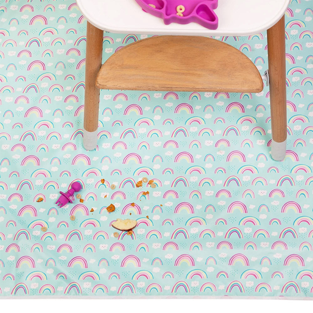 A light blue and rainbow patterned splat mat underneath a white and wooden highchair with food and pink chewtensil fork on the mat 