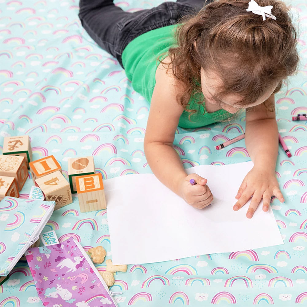 A young child lies on a pastel rainbow-patterned mat, drawing on a white sheet of paper with a purple crayon. Wooden letter blocks, a bumkins zip snack bag, and other toys are scattered around on the splat mat.